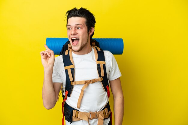 Jovem alpinista russo com uma grande mochila isolada em um fundo amarelo com a intenção de descobrir a solução enquanto levanta um dedo