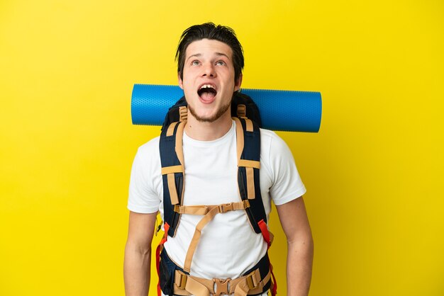 Jovem alpinista russo com uma grande mochila isolada em um fundo amarelo, olhando para cima e com expressão de surpresa