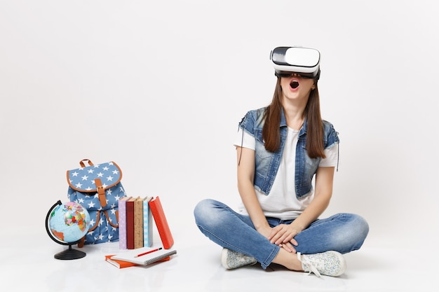 Jovem aluna animada com a boca aberta em óculos de realidade virtual, desfrutando de sentar-se perto da mochila de livros escolares do globo, isolados na parede branca