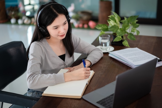 Jovem aluna asiática usa fones de ouvido sem fio, escreva no caderno para estudar idiomas online, assista e ouça o palestrante, webinar via videochamada e-learning em casa, educação à distância