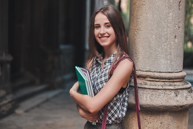 Jovem aluna com cadernos coloridos se apoiando em uma coluna e sorrindo