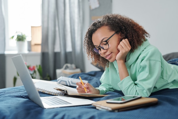 Foto jovem aluna contemporânea em óculos, deitada na cama em frente ao laptop, olhando as anotações enquanto se prepara para os exames