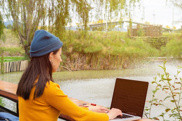 Jovem aluna de costas usando um gorro azul e um suéter amarelo em frente a um lago enquanto usa seu laptop