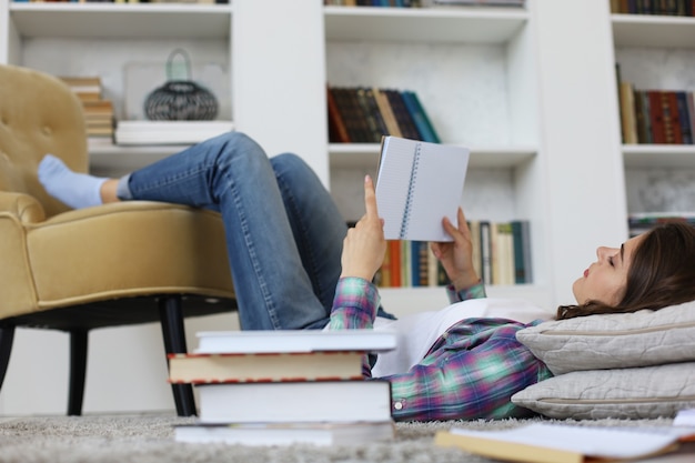 Jovem aluna estudando em casa, preparando-se para os exames universitários, deitada no chão, contra o aconchegante interior doméstico, rodeada de uma pilha de livros.