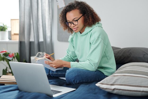 Jovem aluna mestiça em trajes casuais, sentada na cama em frente ao laptop, fazendo anotações e ouvindo a professora durante a aula online