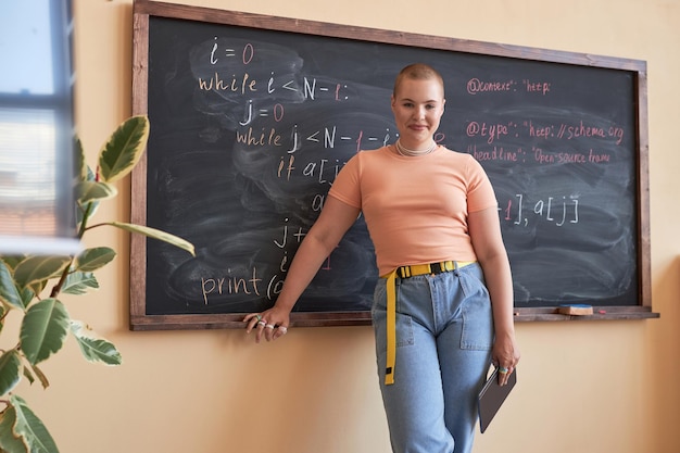 Jovem aluna ou professora de informática bem-sucedida com tablet na mão, parada no quadro-negro em sala de aula e olhando para a câmera