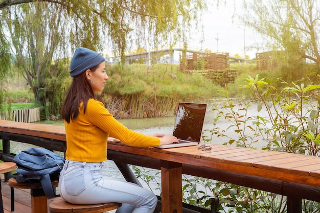 Jovem aluna sentada em um banco de madeira vestindo um gorro azul e um suéter amarelo em frente a um lago enquanto usa seu laptop