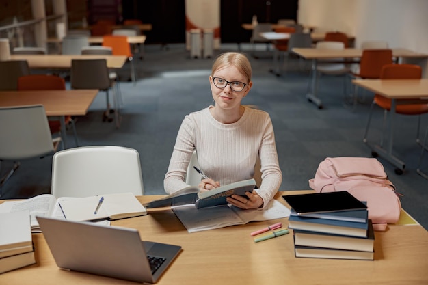 Jovem aluna talentosa estudando na biblioteca da universidade usa laptop e livros