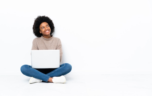 Jovem americana Africano com um laptop sentado no chão olhando para cima enquanto sorrindo