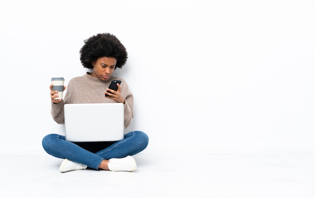 Jovem americana Africano com um laptop sentado no chão segurando café para levar e um celular