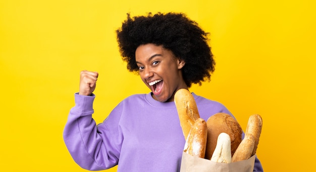 Jovem americana africano comprando algo pão isolado na parede amarela comemorando uma vitória