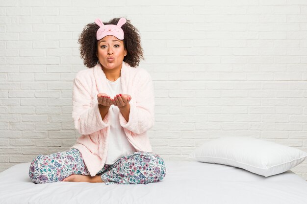 Jovem americana africano na cama vestindo pijama dobrar os lábios e segurando as palmas das mãos para enviar beijo de ar.