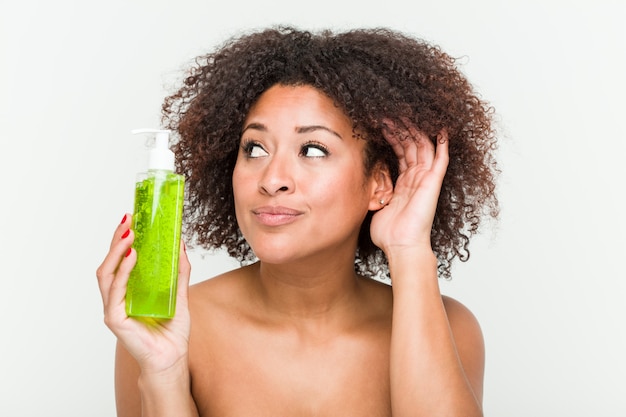 Foto jovem americana africano segurando a garrafa de aloe vera, tentando ouvir uma fofoca.