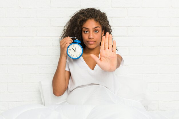 Foto jovem americana africano sentado na cama segurando um despertador em pé com a mão estendida, mostrando o sinal de stop, impedindo-o.