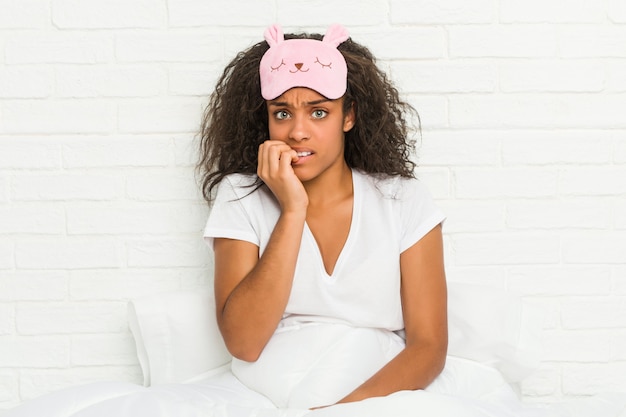 Jovem americana Africano sentado na cama vestindo uma máscara de dormir roer unhas, nervoso e muito ansioso.