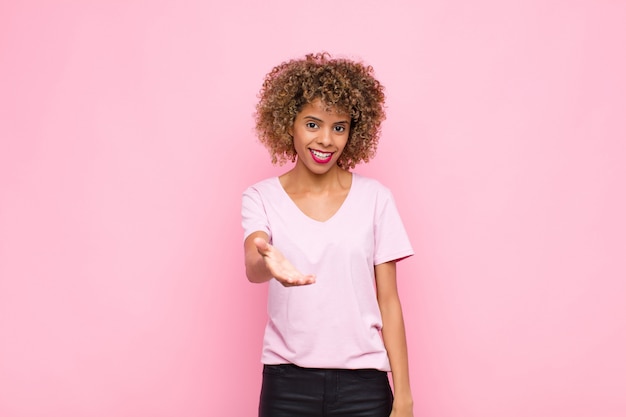 Jovem americana africano sorrindo, olhando feliz, confiante e amigável, oferecendo um aperto de mão para fechar um acordo, cooperando contra a parede rosa