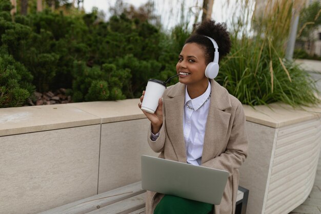 Jovem americana em fones de ouvido com laptop bebendo café lá fora