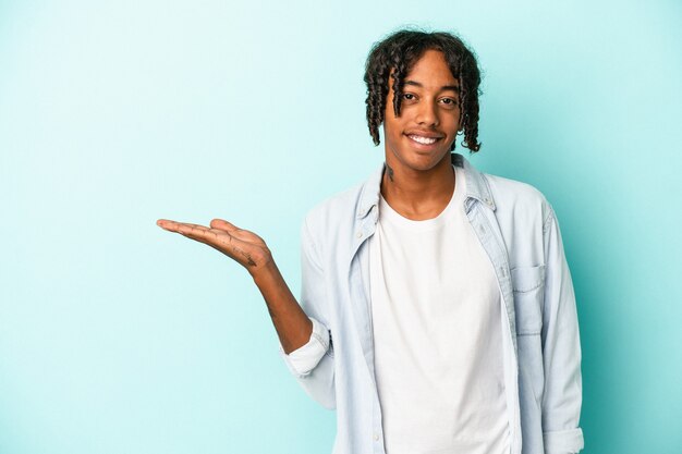 Jovem americano africano isolado em um fundo azul, mostrando um espaço de cópia na palma da mão e segurando a outra mão na cintura.