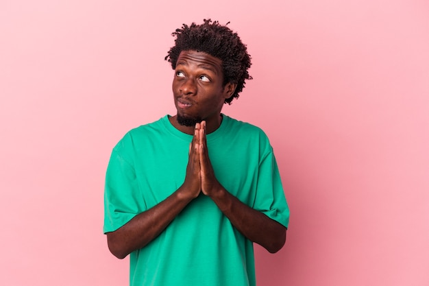 Jovem americano africano isolado em um fundo rosa orando, mostrando devoção, pessoa religiosa em busca de inspiração divina.
