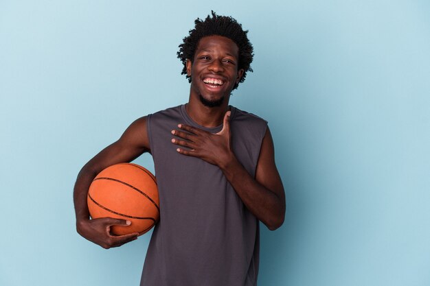 Jovem americano africano jogando basquete isolado no fundo azul ri alto mantendo a mão no peito.