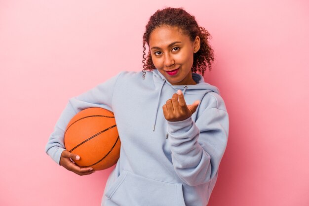 Jovem americano africano jogando basquete isolado no fundo rosa, apontando com o dedo para você, como se fosse um convite para se aproximar.