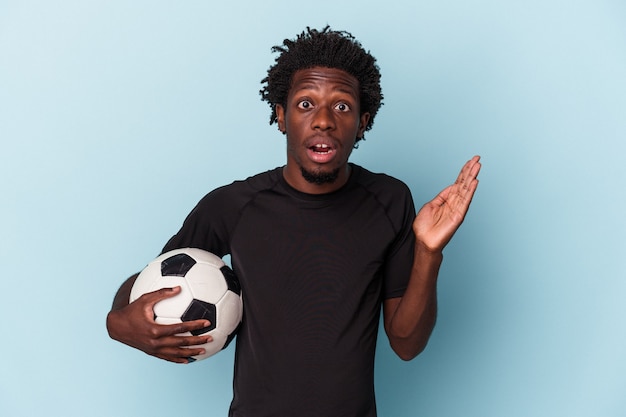 Jovem americano africano jogando futebol isolado em um fundo azul surpreso e chocado.