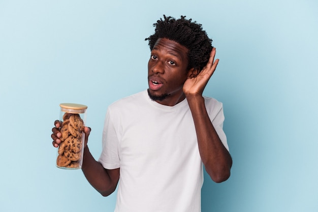 Jovem americano africano segurando biscoitos de chocolate isolados em um fundo azul, tentando ouvir uma fofoca.
