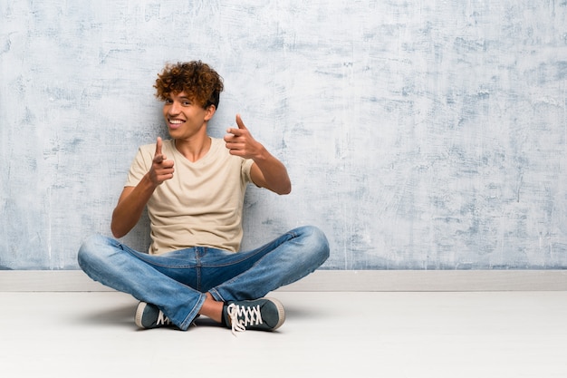 Jovem americano africano sentado no chão, apontando para a frente e sorrindo