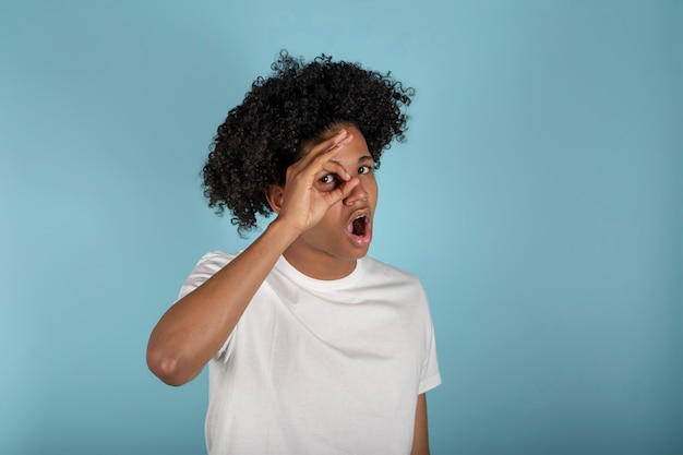 Jovem americano africano vestindo camiseta branca básica sobre fundo azul pastel isolado espreitando em estado de choque, cobrindo o rosto e os olhos com a mão, olhando através dos dedos com uma expressão envergonhada.