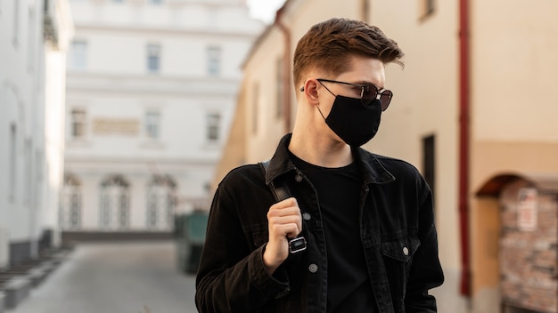 Jovem americano com penteado em óculos de sol da moda na elegante jaqueta jeans preta com elegante máscara preta médica caminha na rua em dia ensolarado.