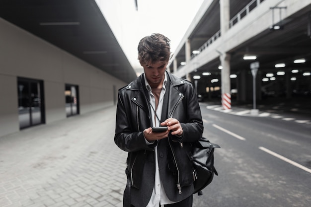 Jovem americano na jaqueta de couro preta com mochila com penteado com telefone celular moderno fica na cidade. cara bonito hipster em roupas da moda fica e olha para o smartphone ao ar livre.