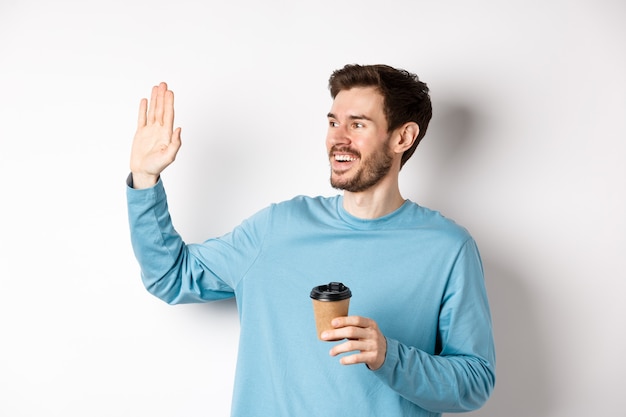 Jovem amigável bebendo café da xícara para viagem e dizendo olá, acenando com a mão para a esquerda e sorrindo, em pé sobre um fundo branco.