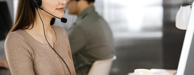 Jovem amigável em fones de ouvido está conversando com o cliente de uma empresa, enquanto está sentado na mesa em um escritório moderno junto com seu colega. Operadores de call center no trabalho.