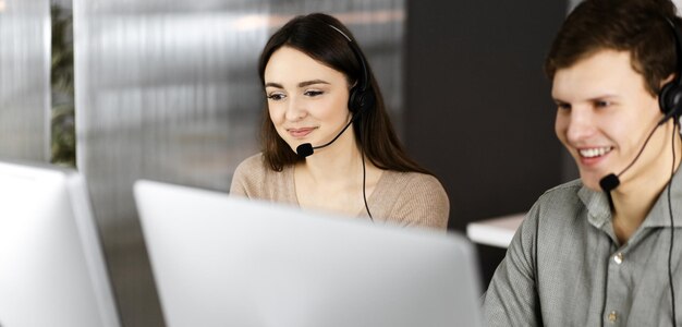 Jovem amigável em fones de ouvido está conversando com o cliente, enquanto está sentado à mesa com uma colega em um escritório. Operadores de call center no trabalho.