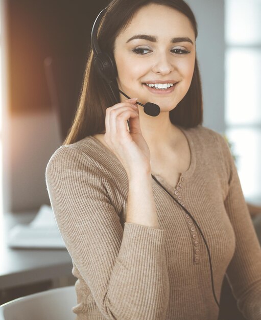 Jovem amigável em fones de ouvido está falando com o cliente de uma empresa, enquanto está sentado na mesa no escritório ensolarado. Operadores de call center no trabalho.