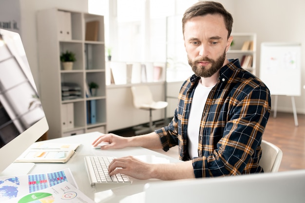 Jovem analista casual sério sentado ao lado do computador, digitando e olhando para a tela do laptop por perto enquanto analisa os dados