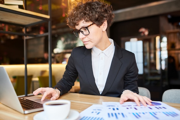 Jovem analista ou corretor elegante, procurando dados on-line enquanto prepara o relatório financeiro no café