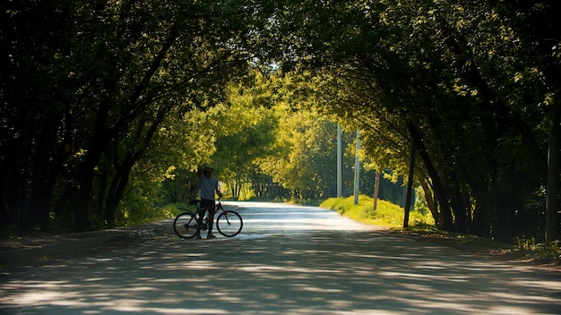 Jovem andando perto de sua bicicleta no arco da árvore do parque verde
