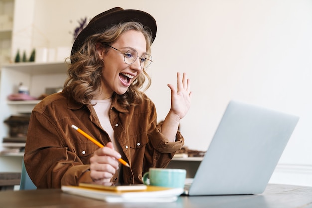 Foto jovem animada com chapéu trabalhando com laptop e acenando com a mão enquanto está sentado à mesa