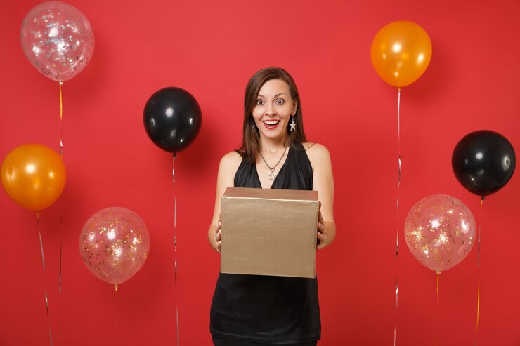Foto jovem animada em um vestido preto, comemorando segurando uma caixa dourada com o presente presente em balões de ar de fundo vermelho brilhante. dia da mulher st., feliz ano novo, conceito de festa de feriado de maquete de aniversário.