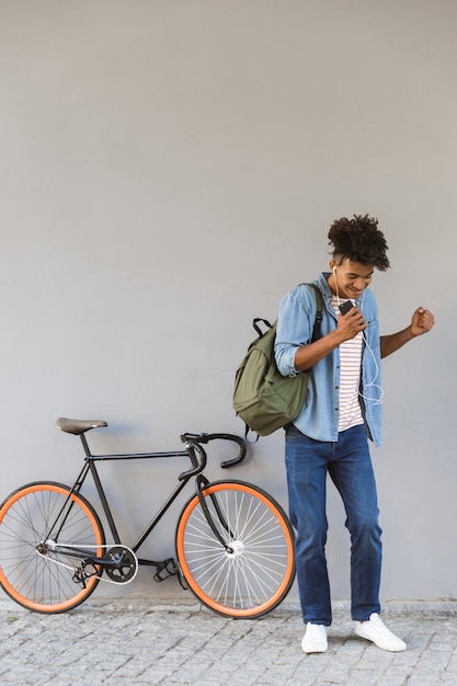 Jovem animado caminhando ao ar livre com uma bicicleta ouvindo música segurando um celular