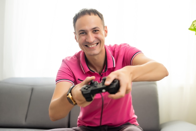 Jovem animado em casa sentado no sofá da sala jogando videogame usando o joystick do controle remoto com expressão facial intensa se divertindo com o vício em jogos