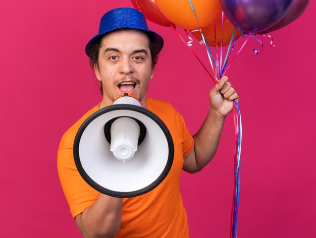Jovem animado usando chapéu de festa segurando balões falando no alto-falante isolado na parede rosa