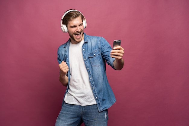 Jovem animado vestindo camisa jeans em pé isolado sobre um fundo rosa, ouvindo música com fones de ouvido e telefone celular