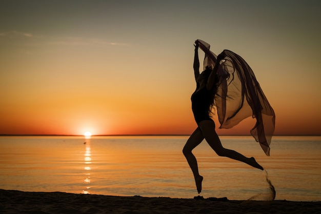 Jovem ao pôr do sol pulando na praia com um pano no fundo do céu