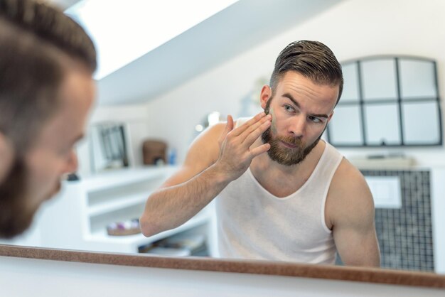 Foto jovem aplicando creme facial enquanto olha para o espelho em casa