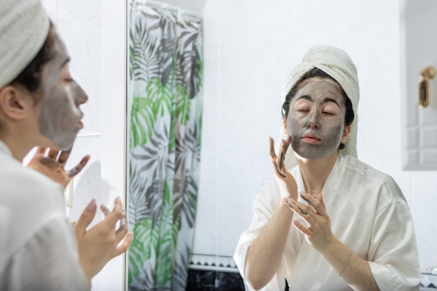 jovem aplicando uma máscara facial de creme preto para a pele em frente ao espelho do banheiro