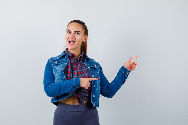 Jovem apontando para o canto superior direito da camisa, jaqueta e parecendo feliz, vista frontal.