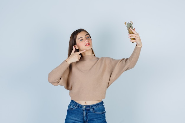 Jovem apontando para o celular de suéter bege, jeans e parecendo confiante. vista frontal.