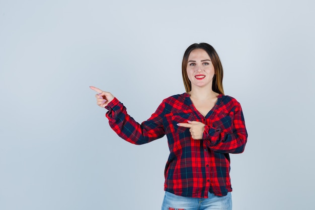 Jovem apontando para o lado esquerdo em camisa quadriculada, jeans e parecendo alegre, vista frontal.